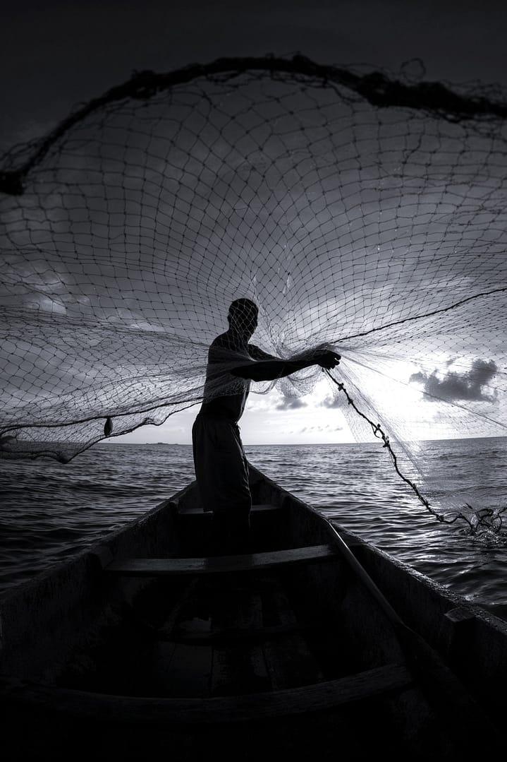 Black and white portrait of a Brazilian fisherman in silhouette casting a large fishing net from a small boat on the water against a dramatic sky with sunlight, titled Coreografia das Águas, from the Cotidiano series in the Brasil Profundo collection, by artist YSA.
