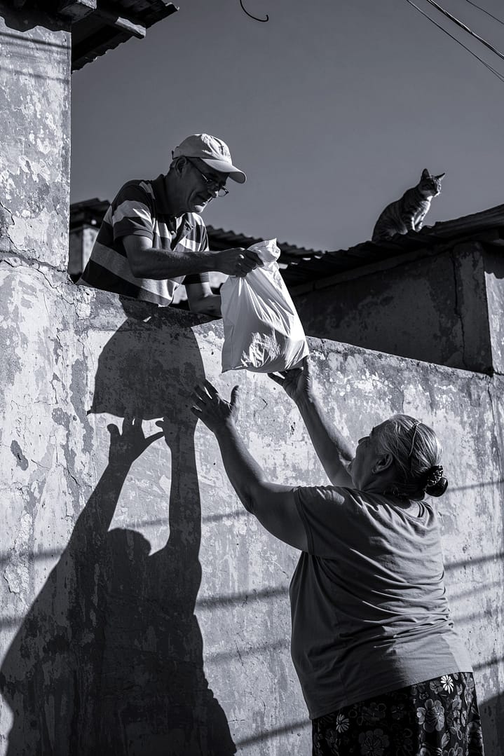 Black and white portrait depicting a woman with outstretched arms passing a package over a rustic wall to a man in a cap and glasses on the other side, with a cat on the roof in the background, titled Isolados na Ilha de Pedra, from the Cotidiano series in the Brasil Profundo collection, by artist YSA.
