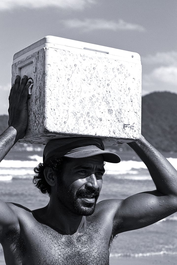 Black and white portrait of a beach vendor carrying a cooler on his head titled João Maré, from the Cotidiano series in the Brasil Profundo collection, by artist YSA.