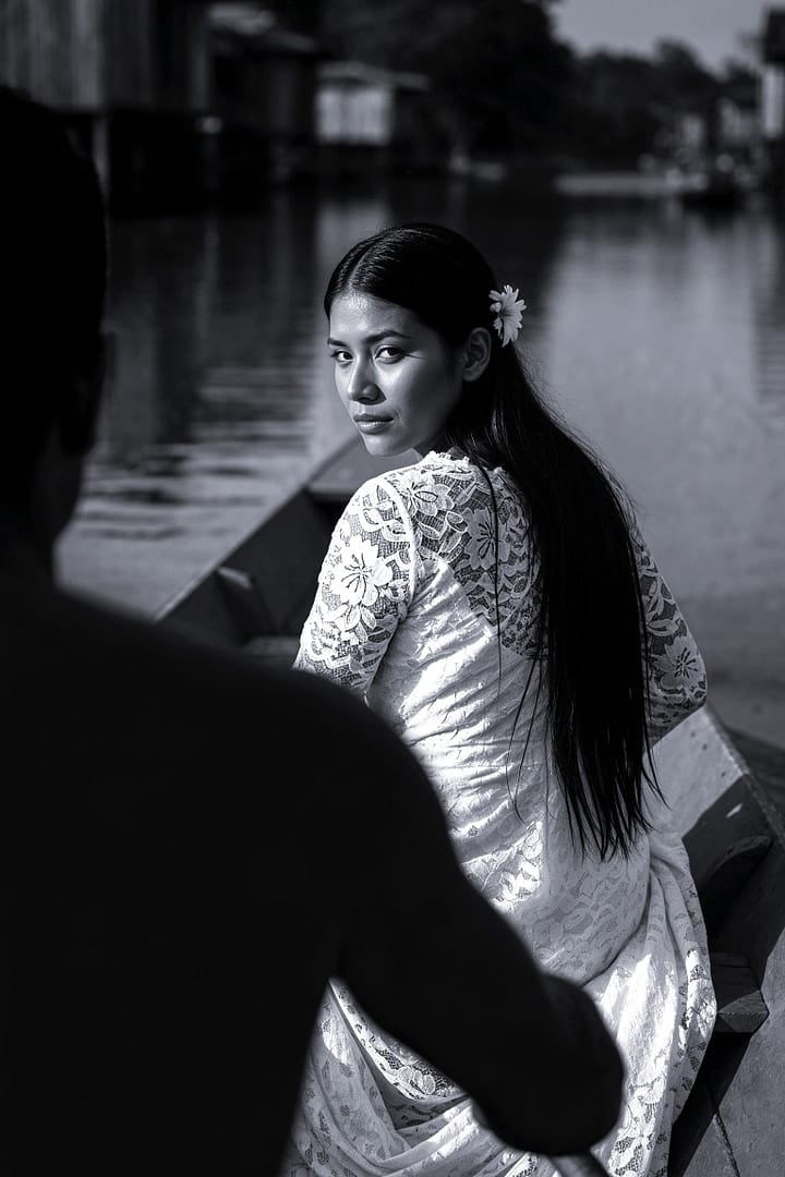 Black and white portrait of a young Brazilian woman with long dark hair and a flower tucked behind her ear, wearing a white lace dress, looking over her shoulder while sitting in a traditional small boat on a river, with the blurred silhouette of a man in the foreground and distant riverine houses in the background, titled A Noiva de Manacapuru, from the Cotidiano series in the Brasil Profundo collection, by artist YSA.
