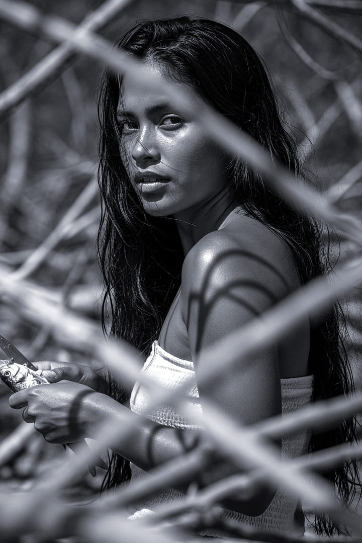 Black and white portrait of a Brazilian woman with long, wet hair, holding and cleaning a fish, surrounded by blurred natural foliage, titled Caiçara, from the Cotidiano series in the Brasil Profundo collection, by artist YSA.