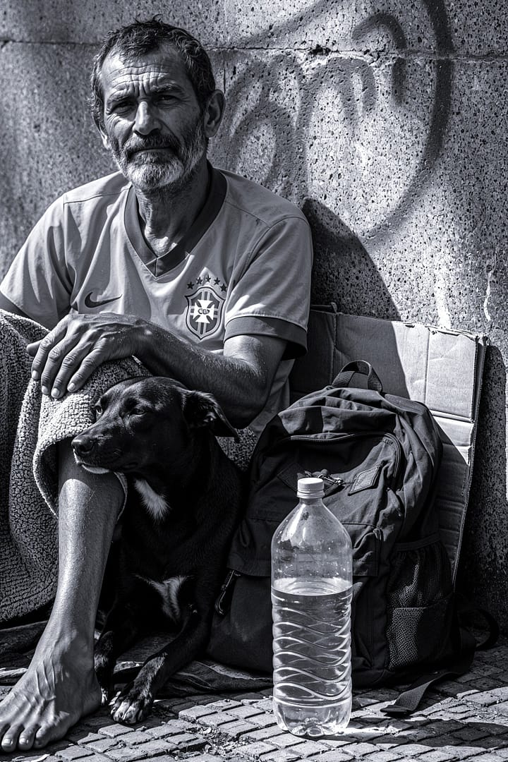 Black and white portrait of a Brazilian man in a Brazil national soccer team jersey, sitting on the ground with a blanket, accompanied by a dog and a bottle of water, against a wall with graffiti in an urban setting, titled Caos e Progresso, from the Cotidiano series in the Brasil Profundo collection, by artist YSA.