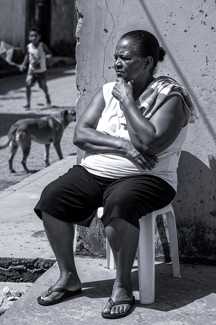 Black and white portrait of a Brazilian woman sitting on a plastic chair in a contemplative pose, with her elbow on her knee and hand on her chin, observing in an outdoor setting with a child and a dog in the blurred background, titled De Sentinela, from the Cotidiano series in the Brasil Profundo collection, by artist YSA.
