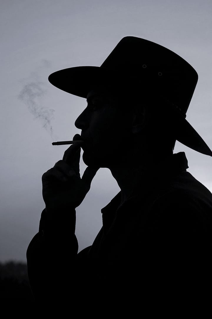 Black and white portrait of a man in silhouette wearing a boiadeiro hat and smoking a cigarette against a bright sky, titled Fogo de Palha, from the Cotidiano series in the Brasil Profundo collection, by artist YSA.