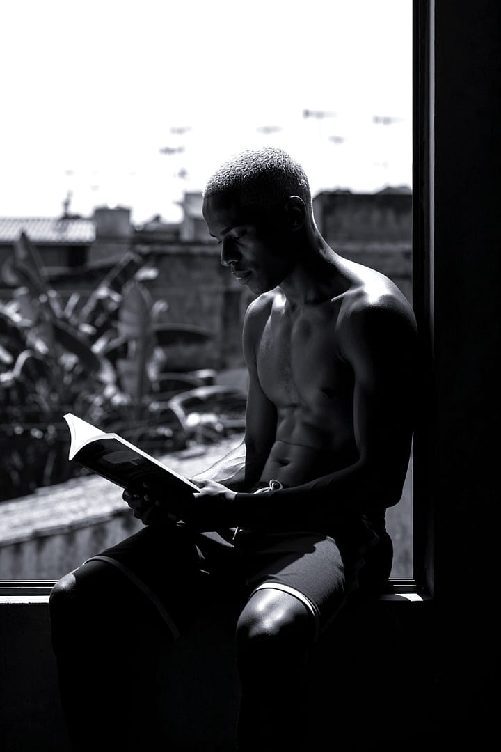 Black and white portrait of a young Black Brazilian man, shirtless, sitting in a window frame and reading a book, silhouetted against a favela background, titled Futuro Emoldurado, from the Cotidiano series in the Brasil Profundo collection, by artist YSA.