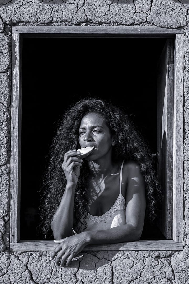 Black and white portrait of a Brazilian woman with long curly hair, eating a slice of mango, looking out from a rustic window with a cracked mud wall, titled Manga Verde com Sal, 2025, from the Cotidiano series in the Brasil Profundo collection, by artist YSA.