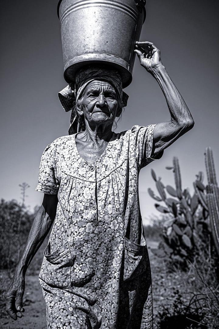 Black and white portrait of an elderly Northeastern Brazilian woman carrying a bucket on her head in the sertão titled Sol de Mandacaru, from the Cotidiano series in the Brasil Profundo collection, by artist YSA.